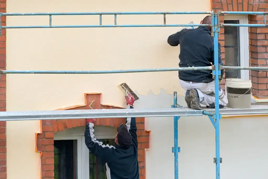 Deux façadiers appliquant un enduit beige sur une maison depuis un échafaudage.
