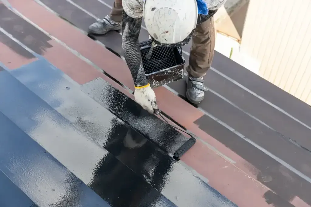 Couvreur appliquant une peinture noire au rouleau sur une couverture métallique.